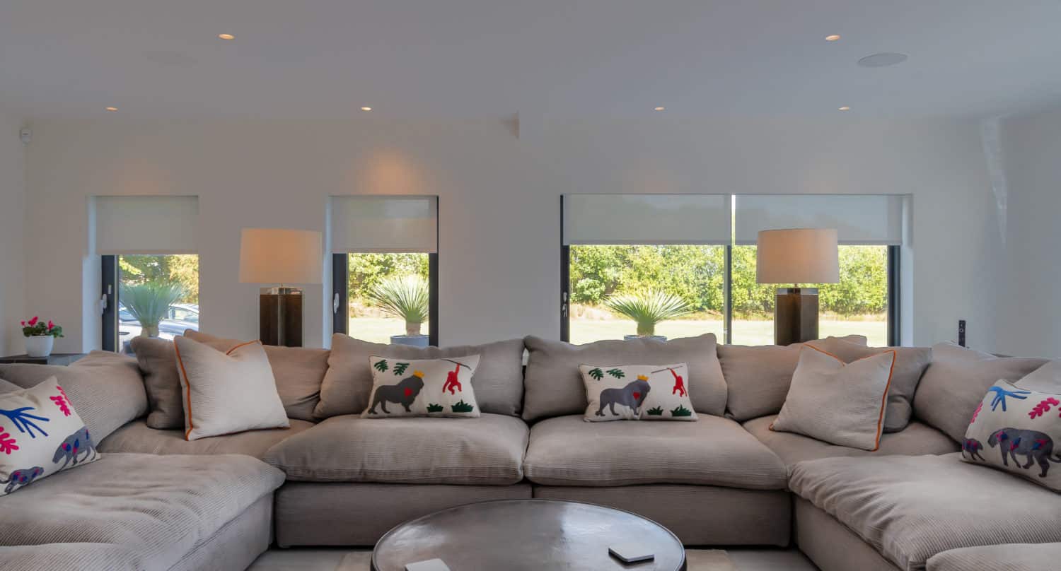 A spacious living room with a large gray sectional sofa decorated with colorful animal-themed pillows, a round coffee table in the center, and two table lamps on either side, featuring sliding doors that open to a lush green outdoor view.