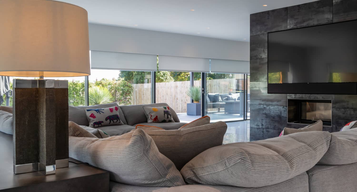 Modern living room with a large gray sectional sofa, colorful animal-print pillows, a table lamp, and a wall-mounted TV above the fireplace. Large windows and bespoke glass solutions offer views of the sunny patio and greenery outside.