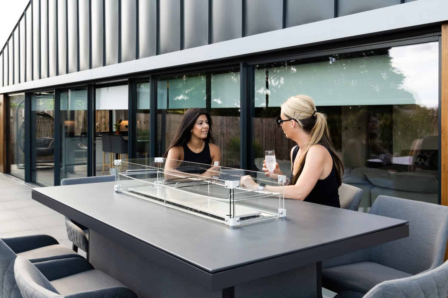 Two women sit at a modern outdoor table, chatting and holding drinks, with bespoke glass solutions showcased in the contemporary glass-walled building and sleek patio furniture in the background.