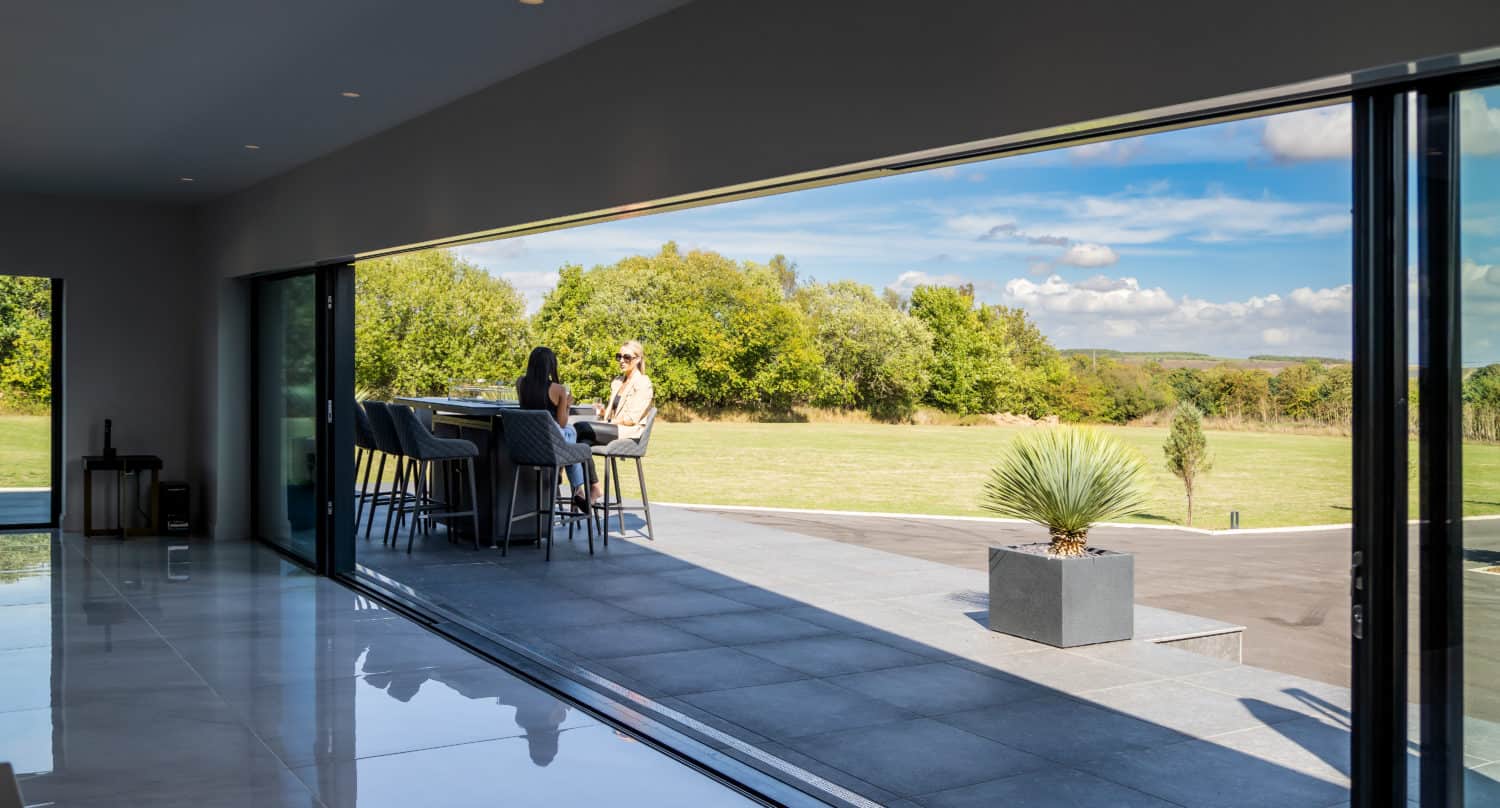 Two people sit at a table on a modern patio outside a house with bespoke glass solutions and large bifold doors, overlooking a grassy lawn and trees under a partly cloudy sky.