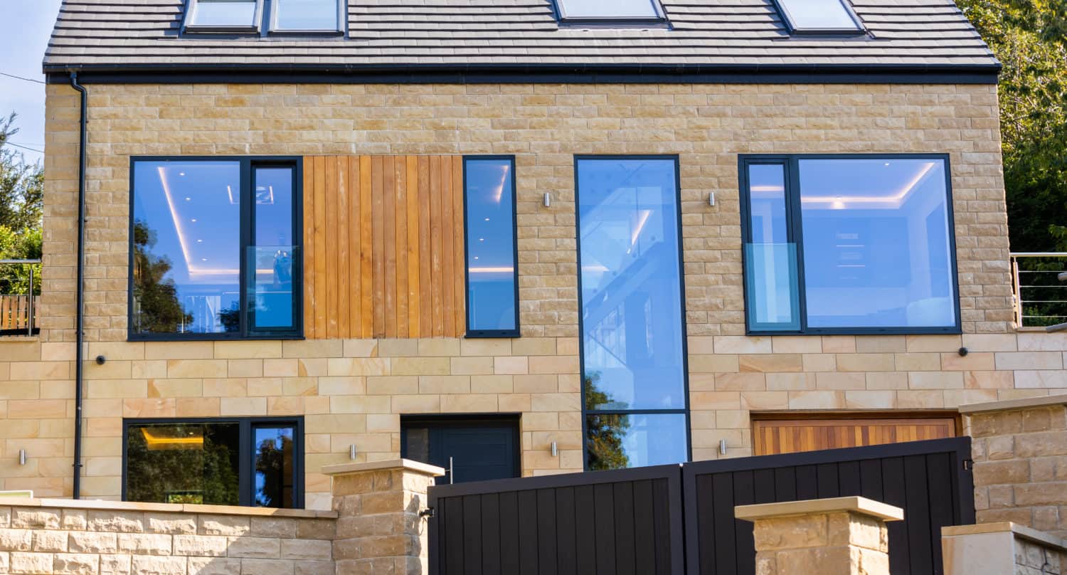 Modern two-story house with large glass windows, bespoke glass solutions, light stone exterior, wooden accents, and a dark sloped roof, behind a stone wall and black gate. Trees are visible in the background.