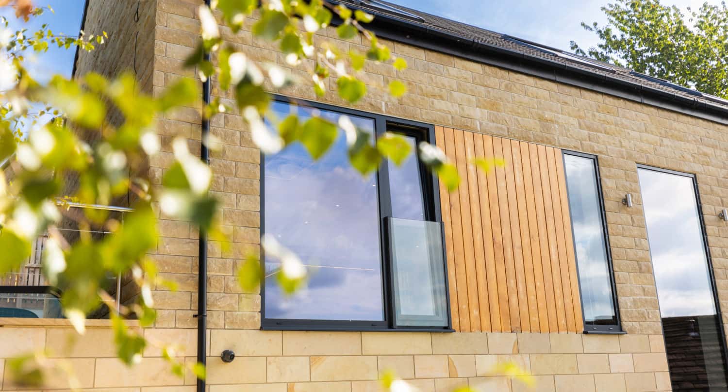 Modern house with large windows, sliding doors, and stone exterior, partially obscured by green leaves in the foreground, under a blue sky with scattered clouds.