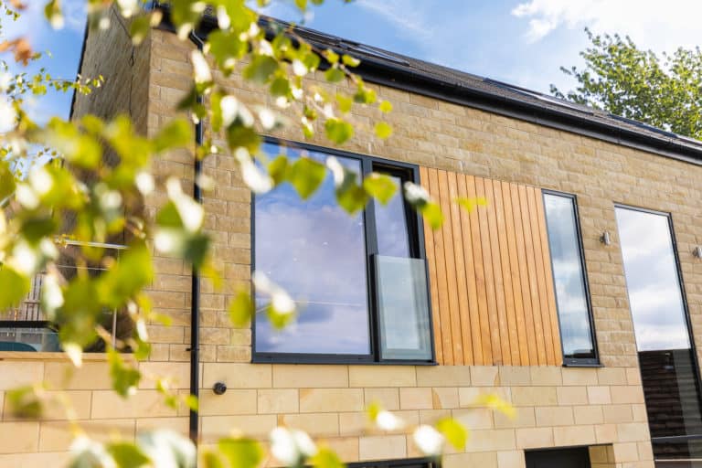Modern house with large windows, sliding doors, and stone exterior, partially obscured by green leaves in the foreground, under a blue sky with scattered clouds.
