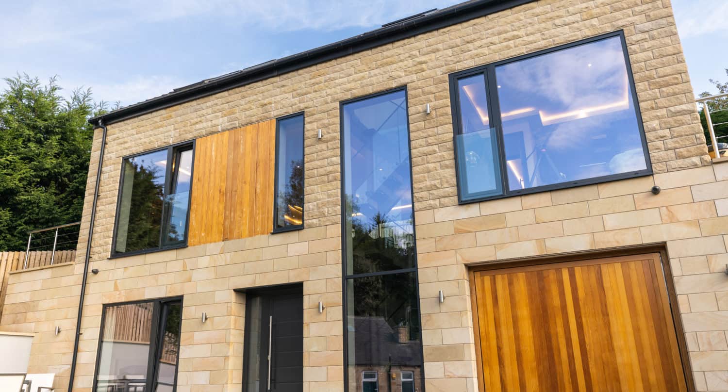 Modern two-story house with large floor-to-ceiling windows, light stone exterior, and wooden doors. The home features clean lines, contemporary design, and sleek sliding doors, with trees visible in the background under a blue sky.