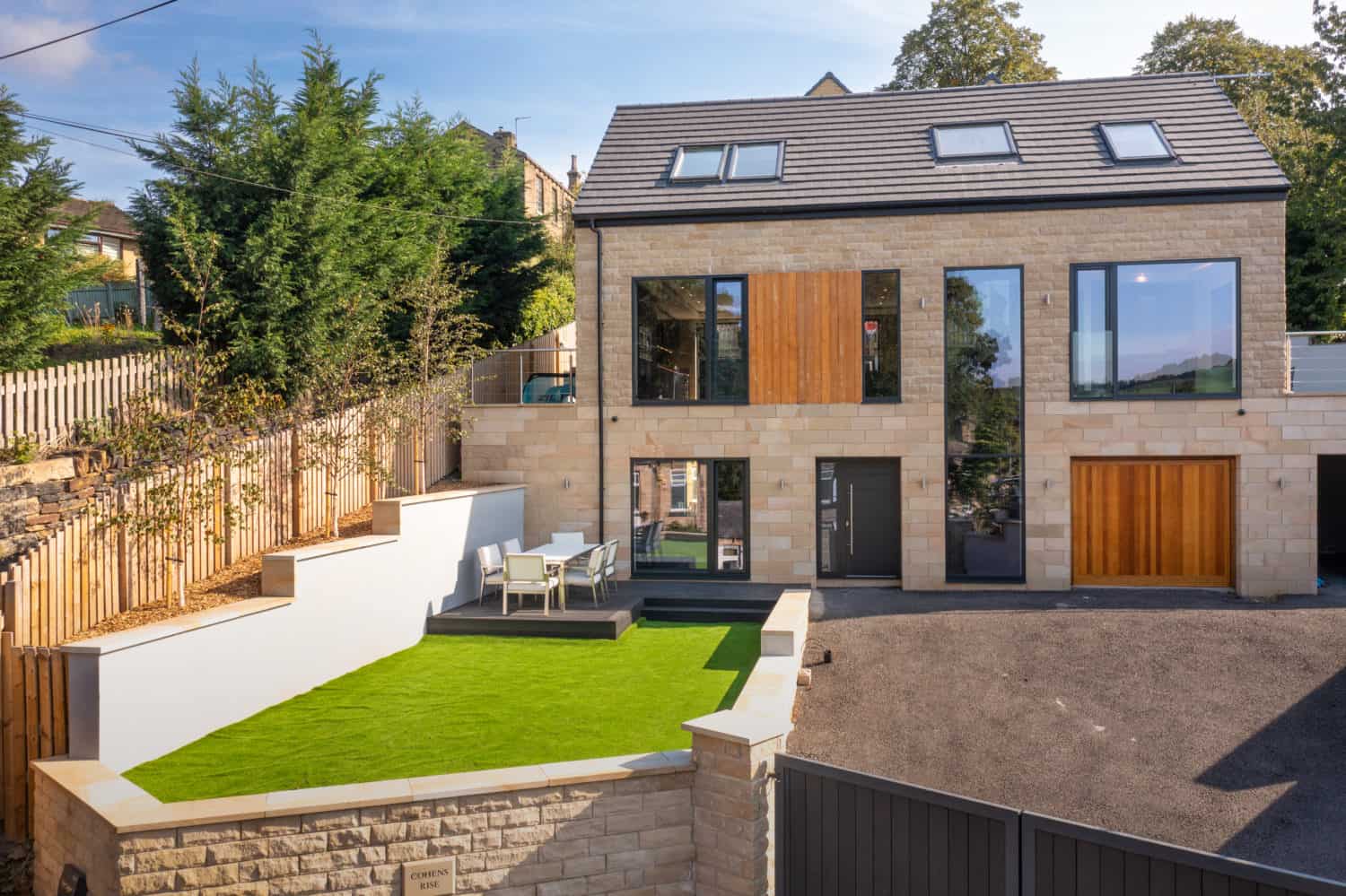 Modern two-story house with large windows, bifold doors, wood accents, and stone exterior. The yard features a walled artificial grass area with outdoor dining furniture, plus a driveway and gated entrance in the foreground.