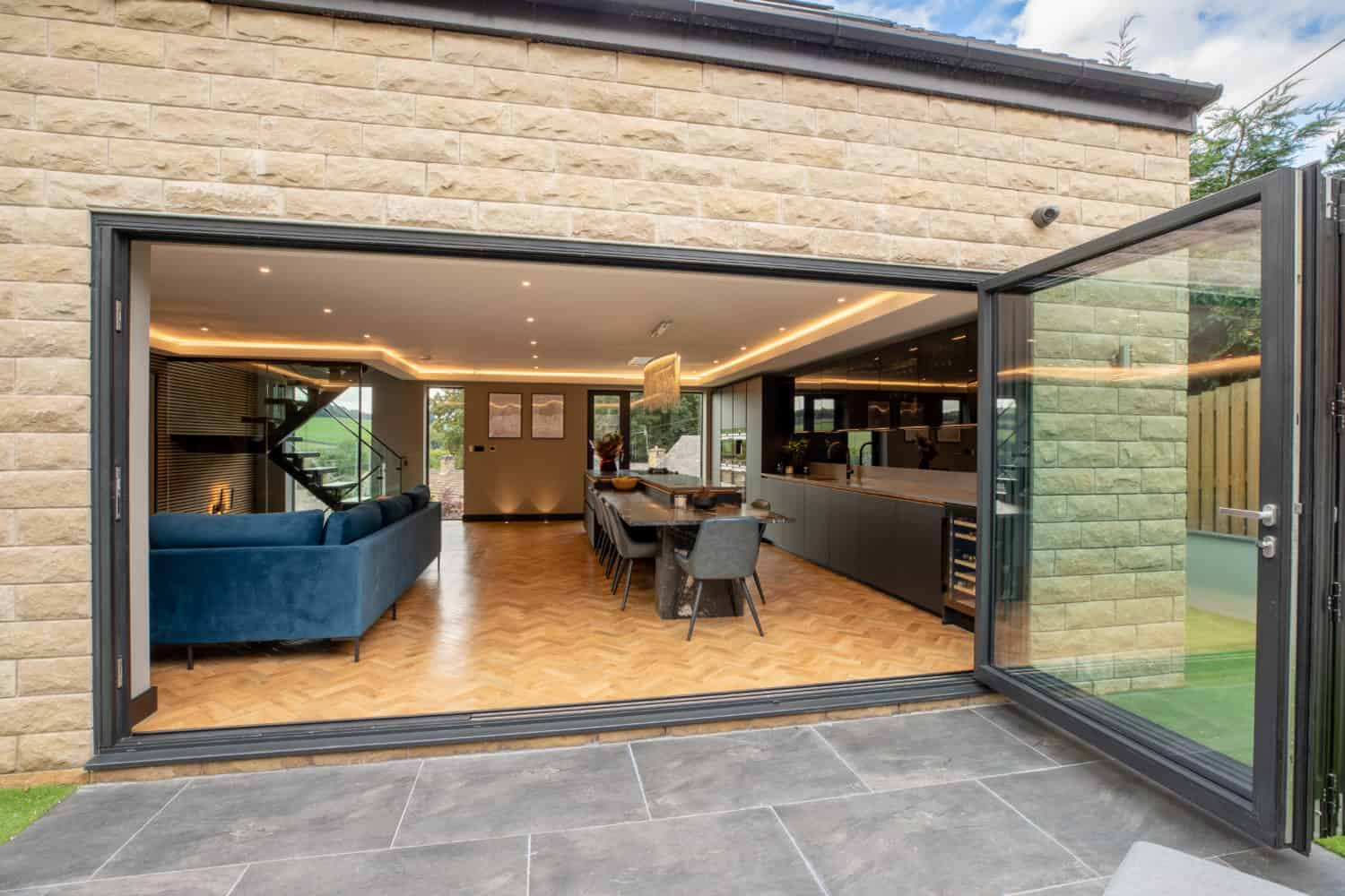 Modern open-plan living and dining area with blue sofa, wooden floor, and bifold doors opening onto a stone patio, seamlessly blending indoor and outdoor spaces.