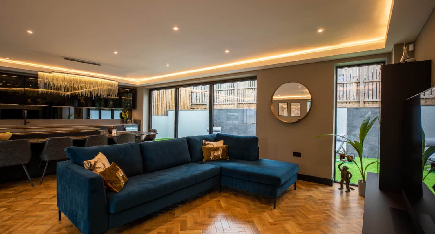 Modern living room with a blue sectional sofa, wood herringbone floor, round mirror, dining area, and large windows letting in natural light. Bespoke glass solutions like glass roofing enhance the airy feel, complemented by warm recessed lighting.