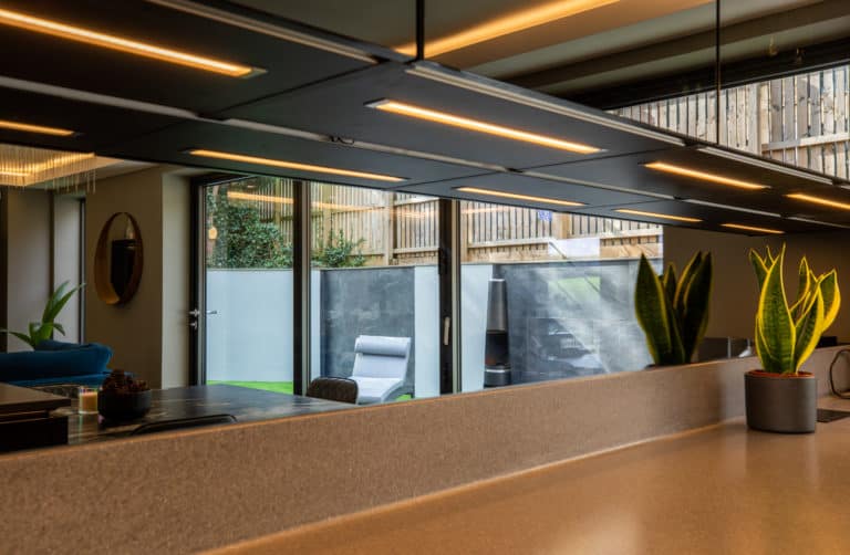 Modern kitchen with a beige countertop, indoor plants, sleek overhead lighting, and large bifold doors leading to an outdoor patio with a lounge chair and greenery outside.