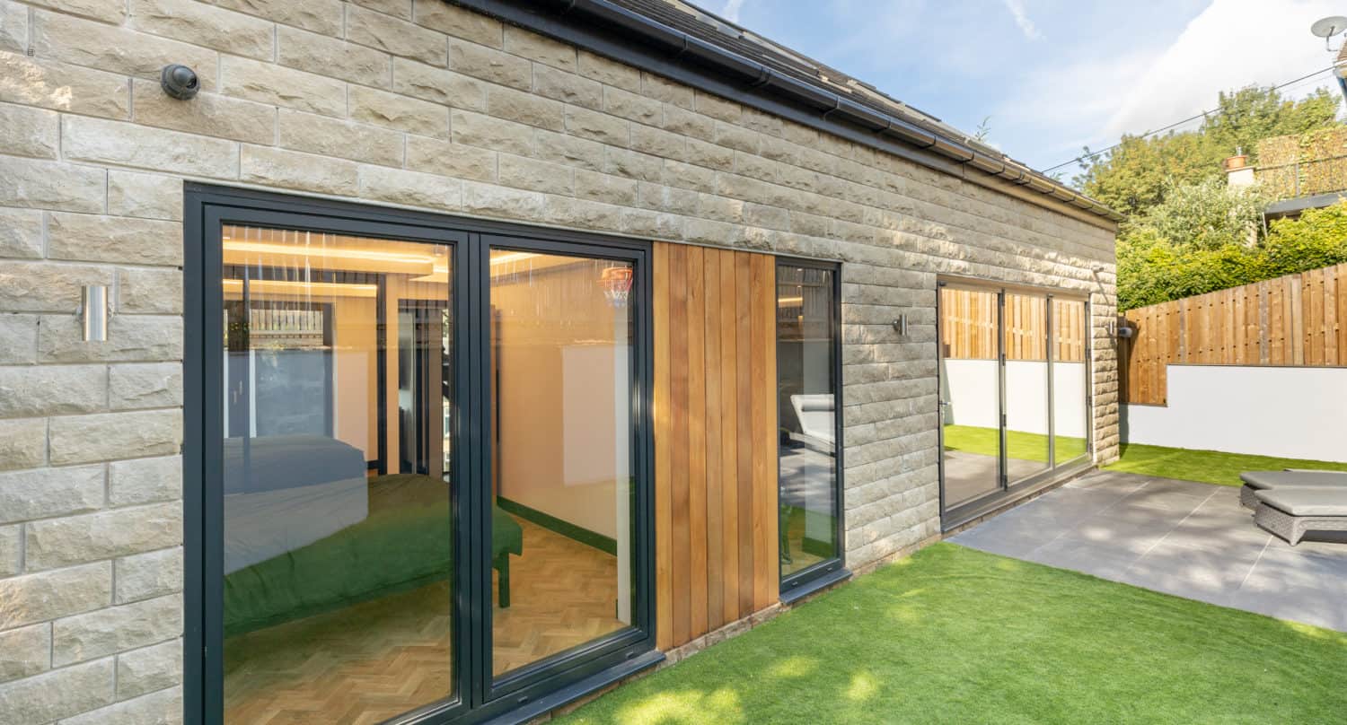 Modern house exterior with stone walls, bespoke glass solutions including large sliding doors, wooden panel accents, and an artificial grass lawn. Bedroom visible through the doors; fenced yard and patio area under a blue sky.