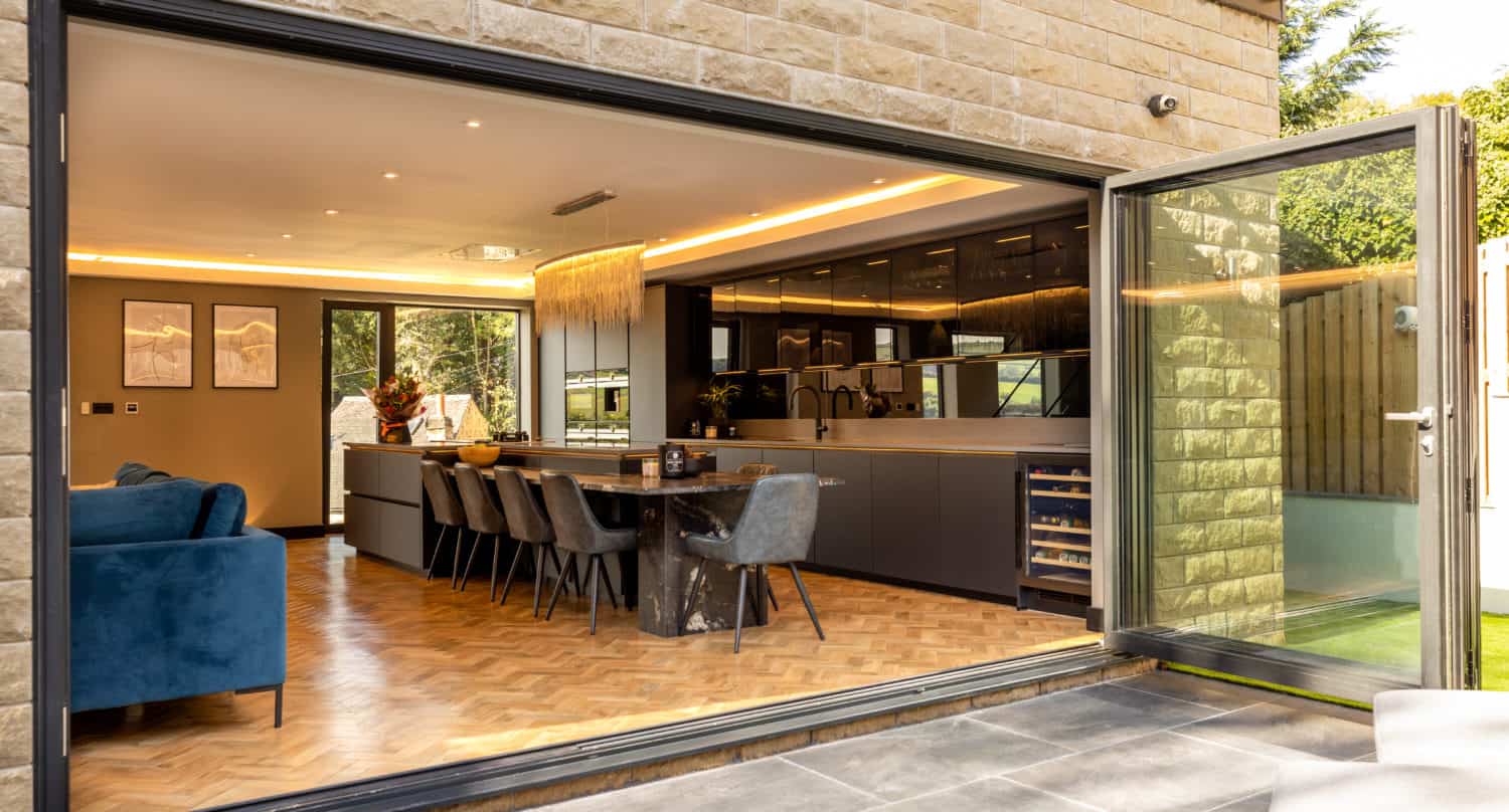 Modern open-plan kitchen and dining area with parquet flooring, visible through large bifold doors opening to a patio. Sleek cabinets, dining table with chairs, and a blue sofa are inside, all enhanced by bespoke glass solutions.