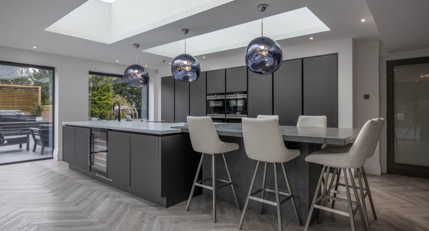 Modern kitchen with dark grey cabinets, a large island with grey countertops, four beige bar stools, and three spherical pendant lights. Skylights, bifold doors, and large windows provide ample natural light. Herringbone wood flooring throughout.
