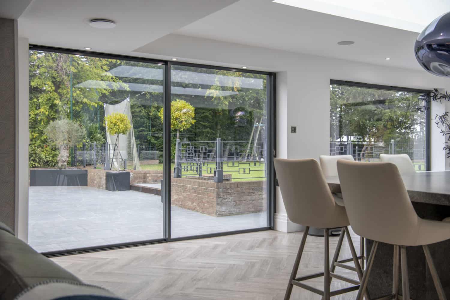 Modern dining area with tall white chairs beside sleek sliding doors, offering a view of a green garden with brick planters, outdoor seating, and trees. Light wood flooring enhances the indoor space.
