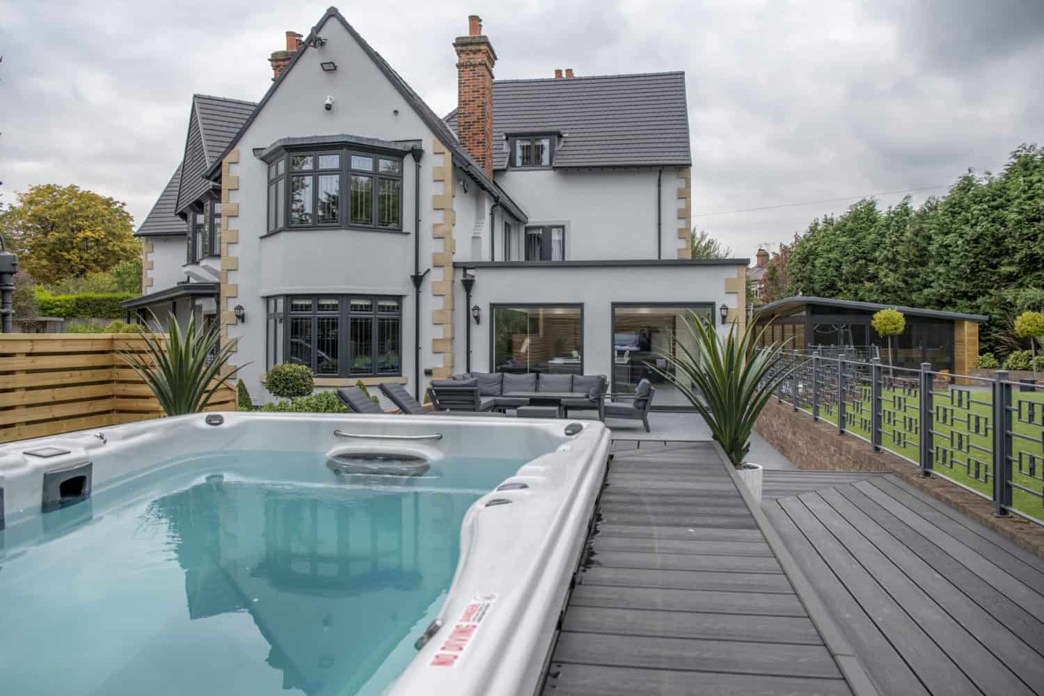 Modern white house with black trim, large windows, and a spacious patio area with outdoor furniture. Sliding doors open to a deck with a hot tub in the foreground, surrounded by greenery and a fenced yard.