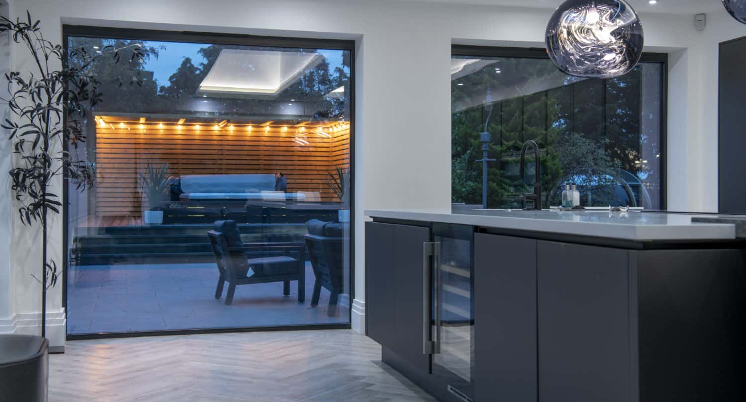 Modern kitchen with dark cabinets, an island, and pendant lights, featuring large windows with bespoke glass solutions that showcase a patio with a covered pool, outdoor seating, and warm accent lighting in the evening.