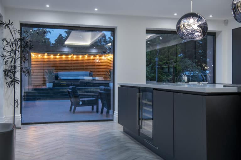 Modern kitchen with dark cabinets, an island, and pendant lights, featuring large windows with bespoke glass solutions that showcase a patio with a covered pool, outdoor seating, and warm accent lighting in the evening.