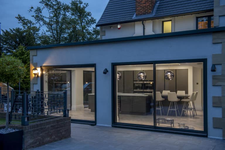 Modern house exterior at dusk with large bifold doors revealing a sleek, contemporary kitchen and dining area inside. Warm indoor lighting contrasts with the blue twilight outside.