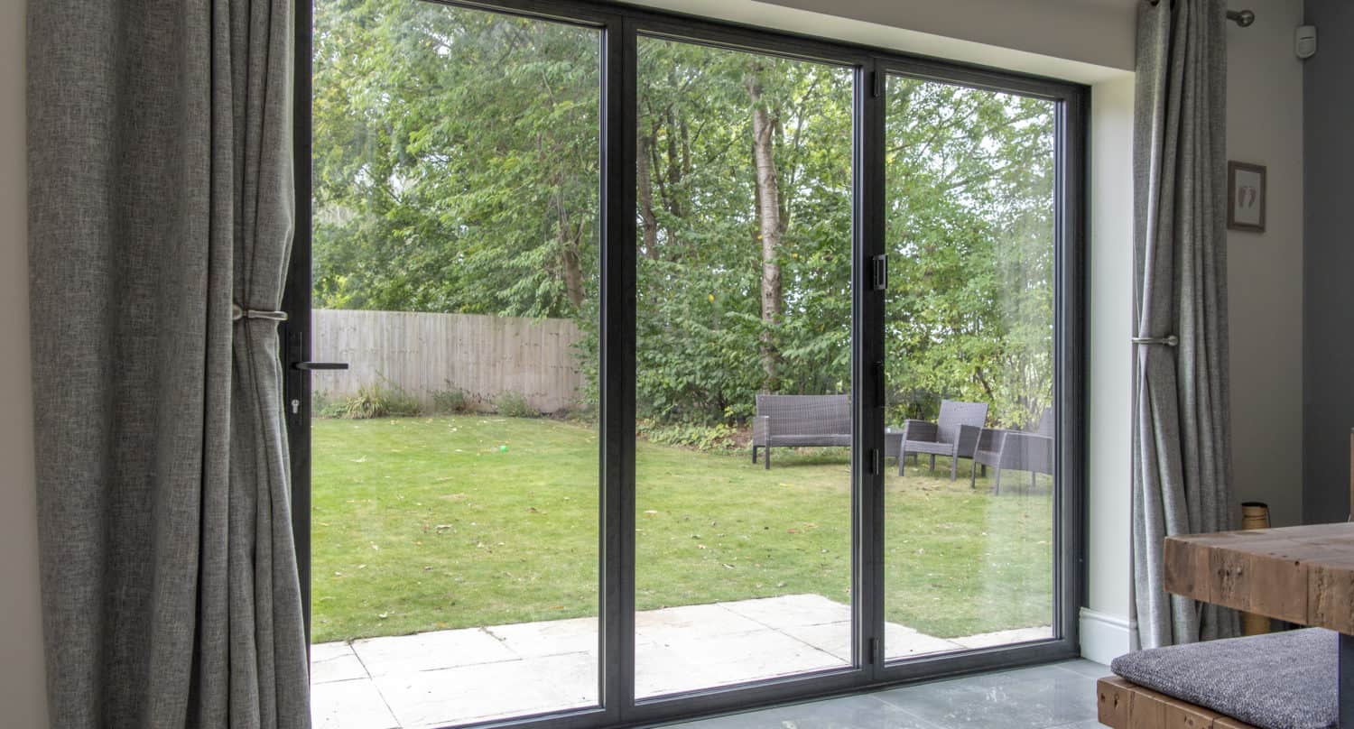A large glass sliding door, crafted with bespoke glass solutions, opens to a backyard with green grass, trees, a wooden fence, and outdoor seating. Gray curtains and a rustic wooden table are visible inside.