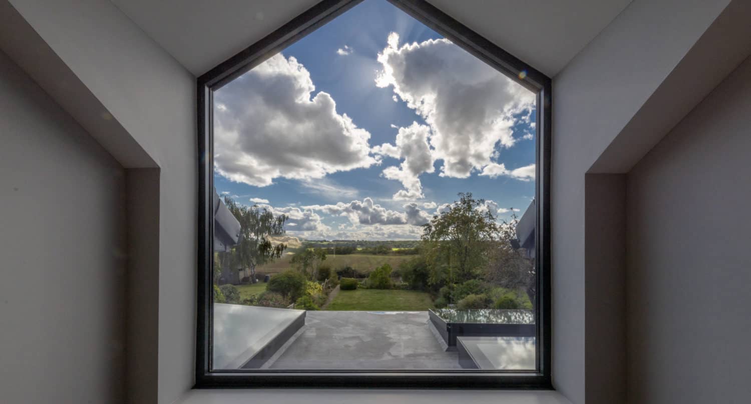 A large, house-shaped window frames a scenic view of a garden with trees and an expansive blue sky. The window’s geometric frame contrasts with the landscape outside, showcasing how bespoke glass solutions blend modern style with nature.