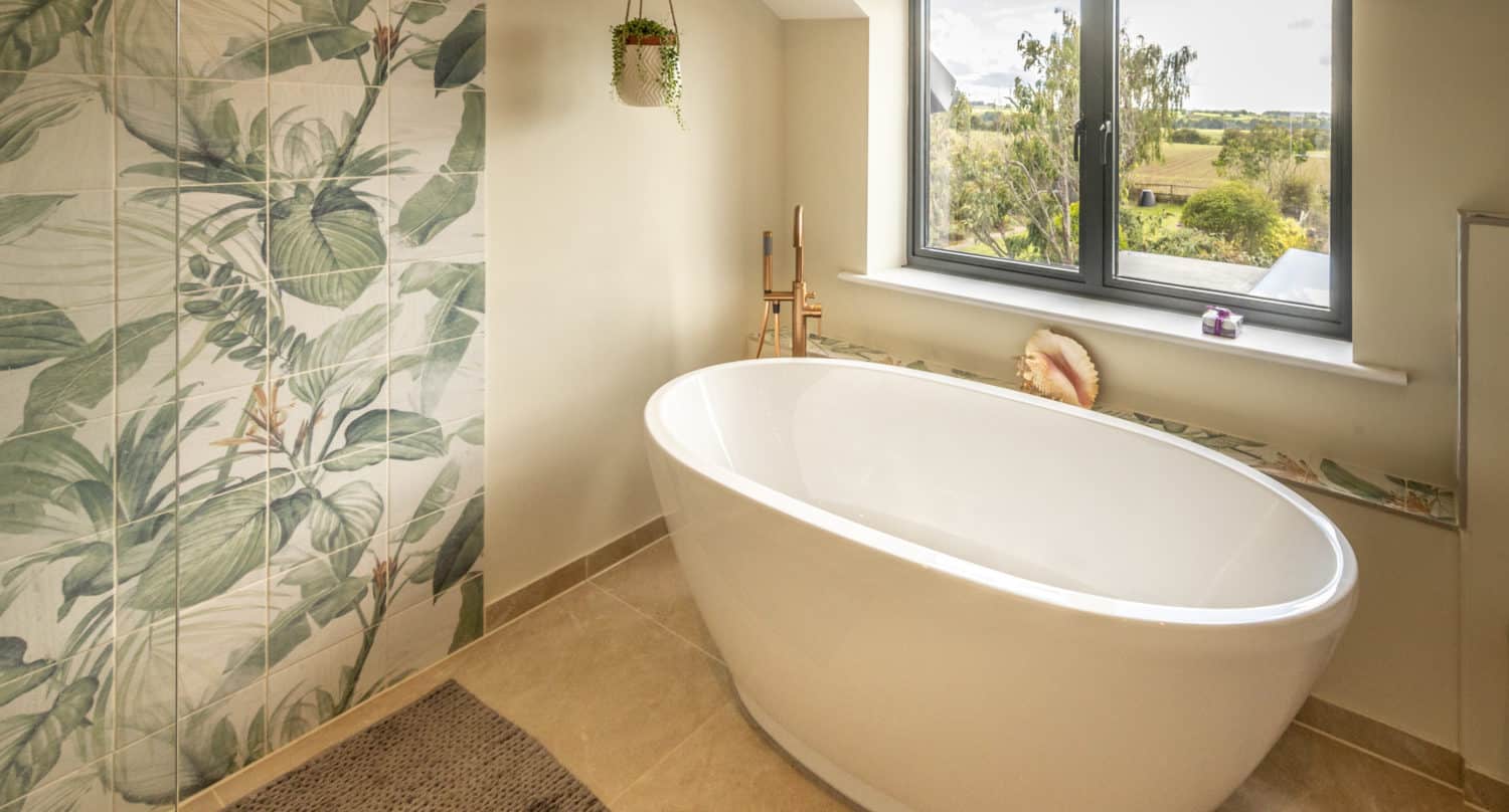 Modern bathroom with a large white oval bathtub, tropical leaf-patterned accent wall, hanging plant, brown mat, and bespoke glass solutions on the window overlooking greenery, creating a bright and serene atmosphere.