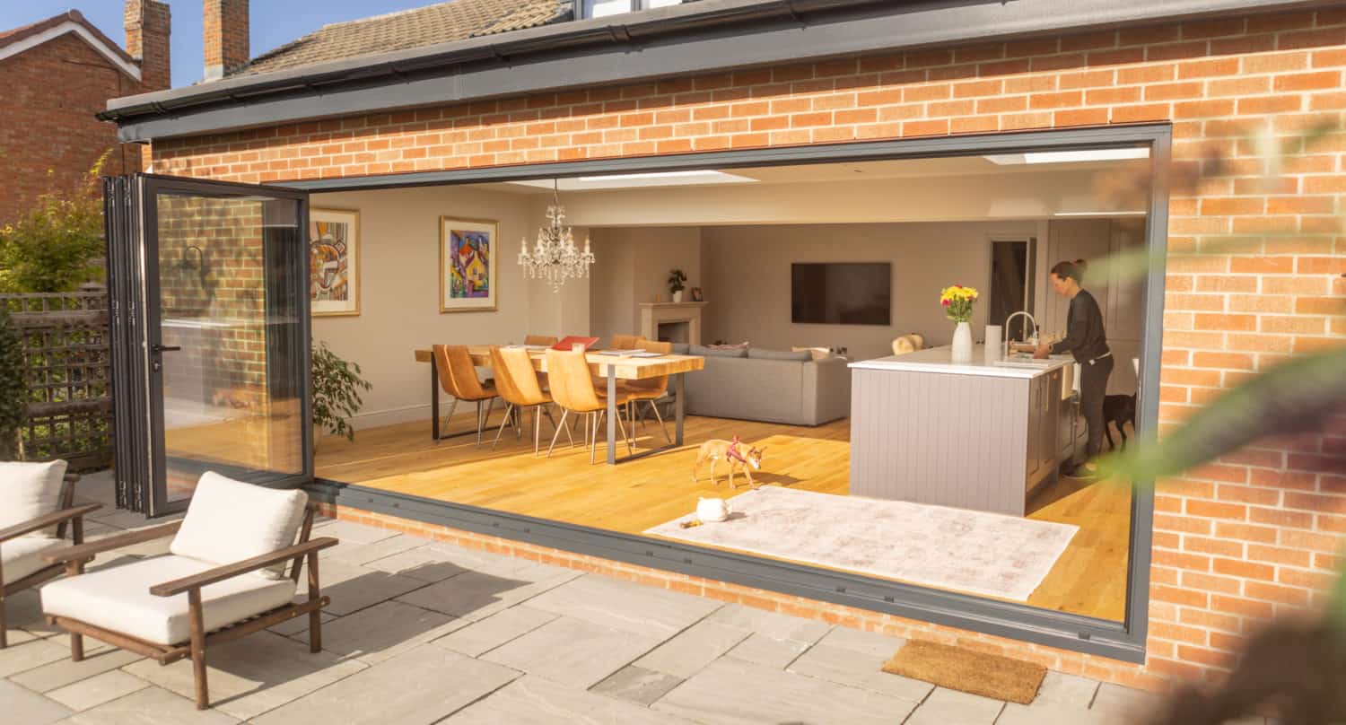 A modern home with bespoke glass solutions features a large open sliding glass door revealing a bright kitchen and dining area. Sunlight fills the space as a woman stands at the island, while two patio chairs sit outside on a stone patio.