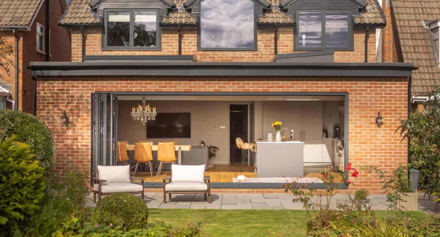 A modern brick house with bespoke glass solutions, featuring large sliding doors that fully open to reveal a spacious kitchen and dining area. The backyard offers a patio with lounge chairs, green lawn, and well-maintained shrubs and plants.