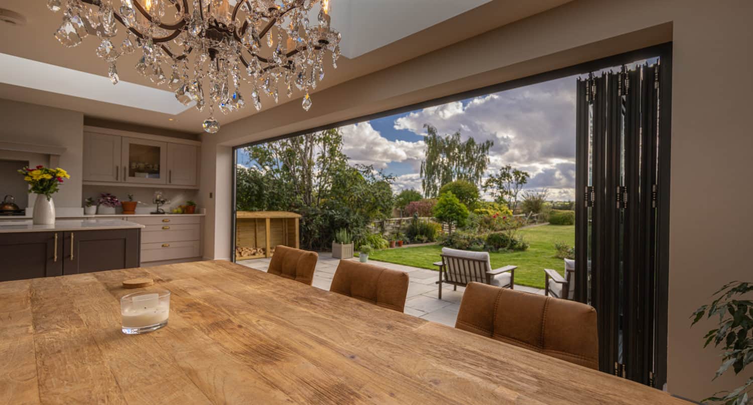 A rustic wooden dining table with brown chairs sits under a crystal chandelier in a kitchen. Large sliding doors open to a lush garden with patio seating, filling the space with natural light and views of greenery and a cloudy sky.