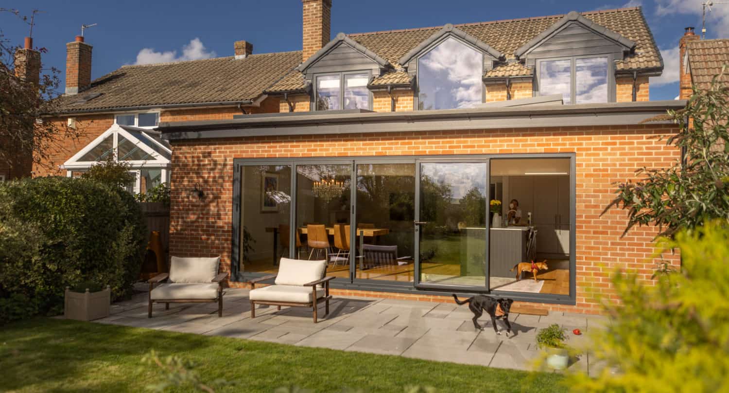A brick house with bespoke glass solutions features large glass doors opening onto a patio with two chairs. A black dog stands outside, while a person is visible inside near the dining table. The garden has green grass and lush plants.