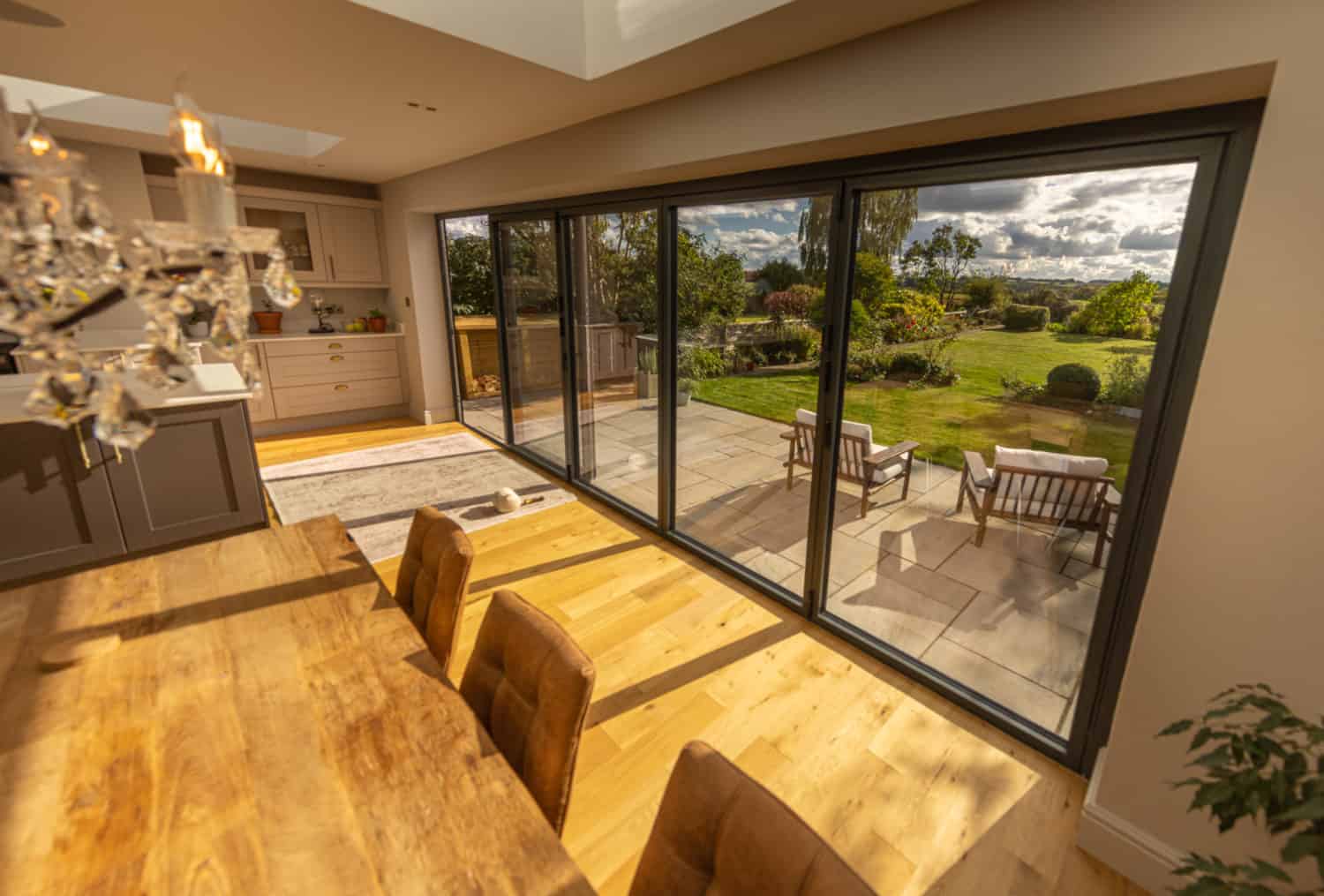 A modern dining area with a wooden table and chairs sits beside large bifold doors that open to a sunny patio with outdoor seating and a landscaped garden beyond.