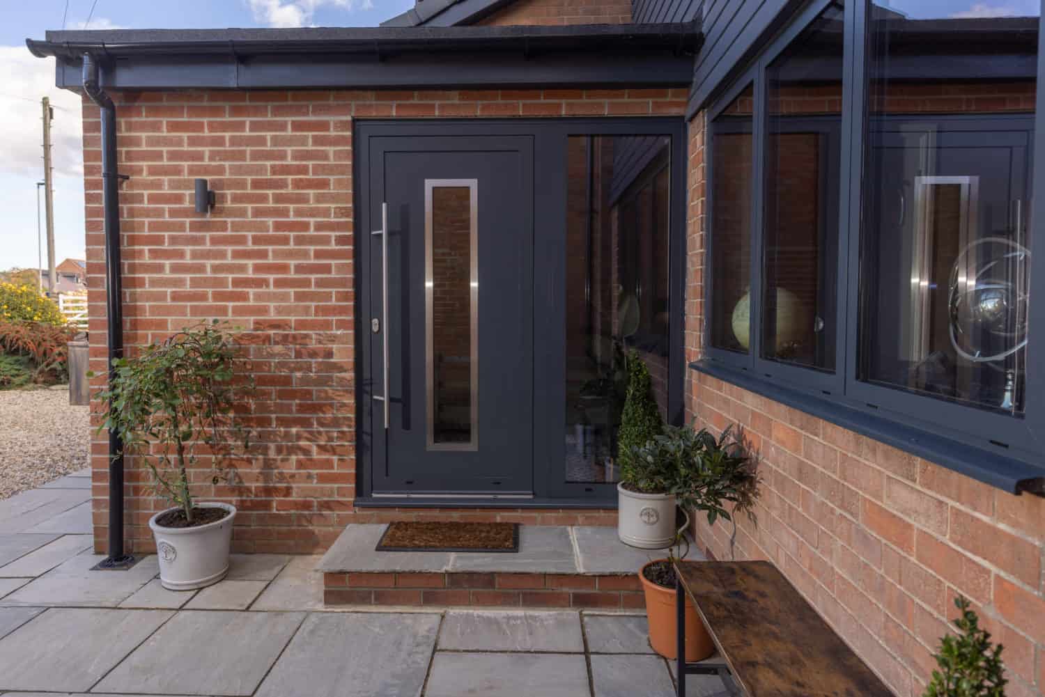 Modern brick house entrance with a dark gray front door featuring a vertical glass panel, flanked by potted plants on a tiled porch and a bench along the wall, complemented by large windows and stylish front doors.