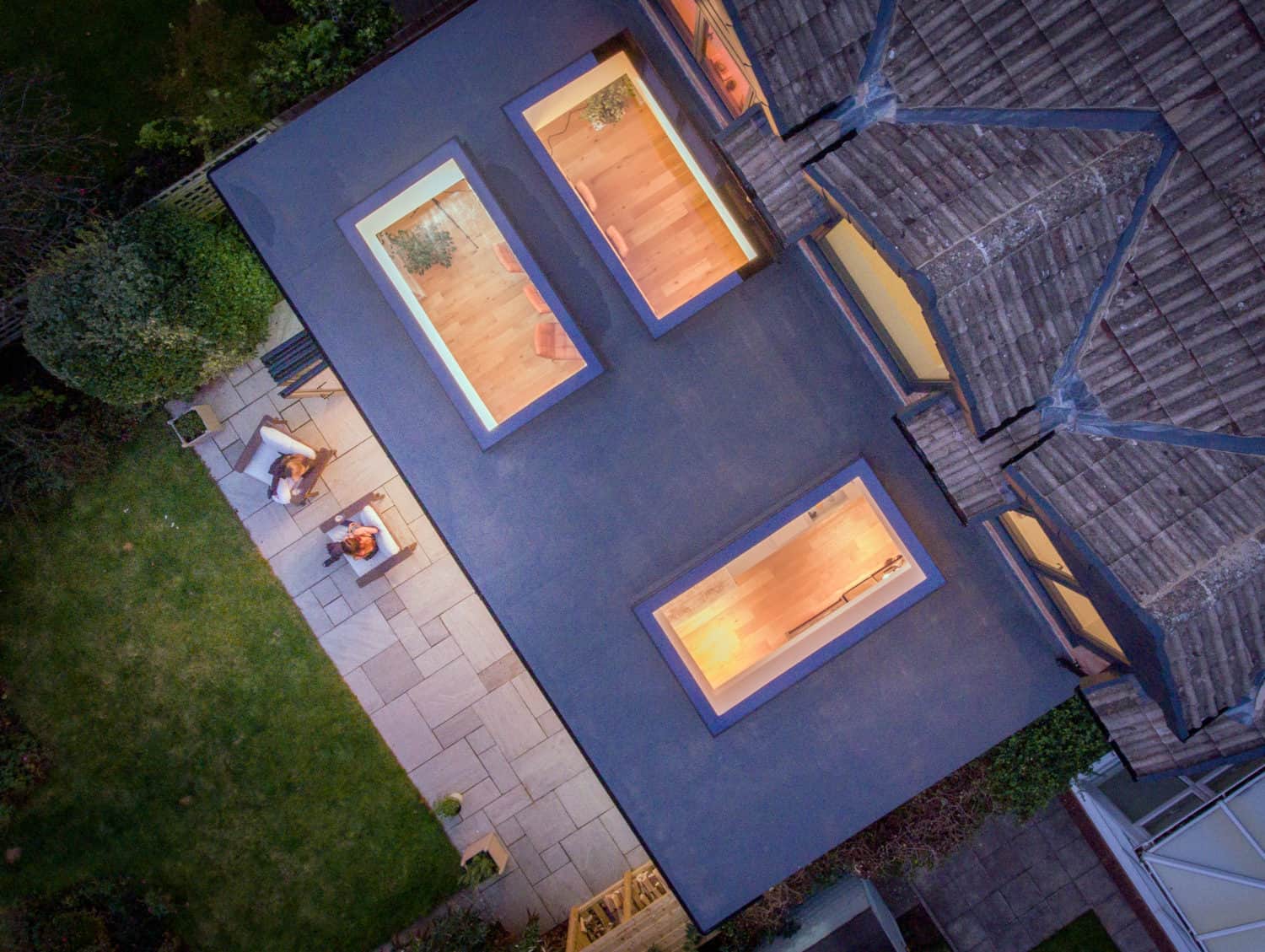 Aerial view of a modern house with bespoke glass solutions, including large skylights on a flat roof, warm lighting inside, and two people sitting at a patio table on a stone terrace beside a green lawn and surrounding garden.