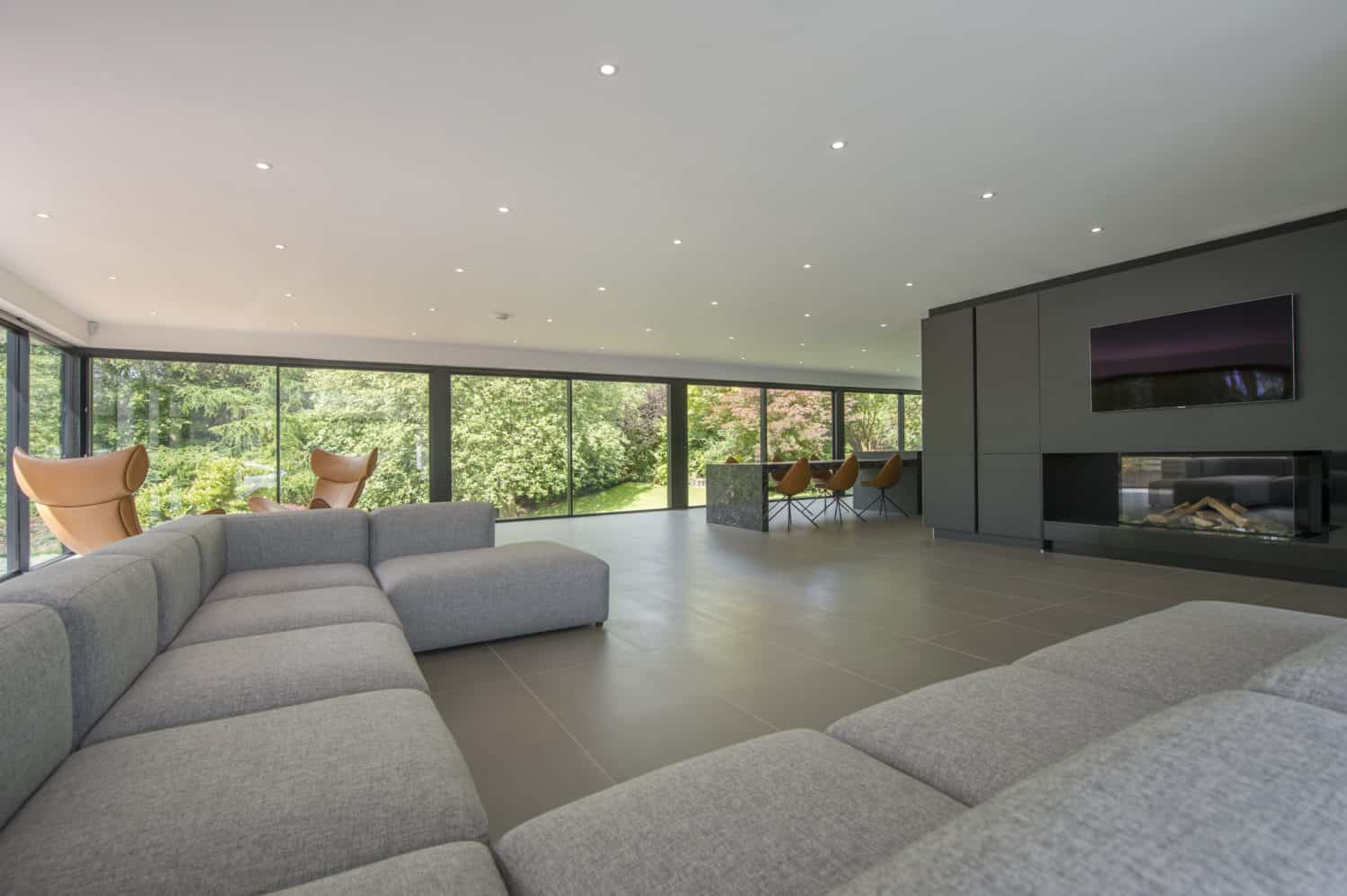 Spacious modern living room with large gray sectional sofa, floor-to-ceiling windows and bifold doors revealing lush greenery outside, a fireplace, wall-mounted TV, and an open dining area with orange chairs.