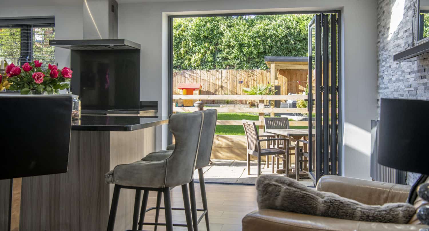 Modern kitchen and living area with bifold doors opening to a sunny patio and garden. Indoor bar stools face a kitchen island, while the backyard—with outdoor table and chairs—blends seamlessly with the bright space.