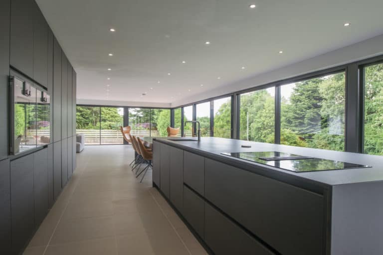 Modern kitchen with dark cabinetry and a large island, featuring an electric cooktop. Bespoke glass solutions, including floor-to-ceiling windows, offer views of green trees and abundant natural light. Dining chairs are arranged near the windows.