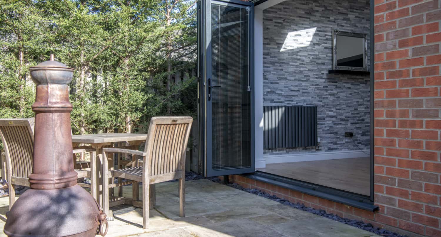 A patio with a wooden table and chairs sits next to a metal chiminea. Large bifold doors open into a room with a gray stone accent wall and radiator. Tall green bushes provide privacy in the outdoor space.