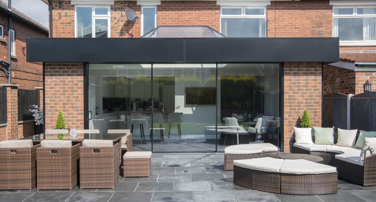 Modern brick house with large glass sliding doors opening to a patio with wicker outdoor furniture, including tables, chairs, and sofas on dark stone tiles, overlooking a green lawn and shaded by partial glass roofing.