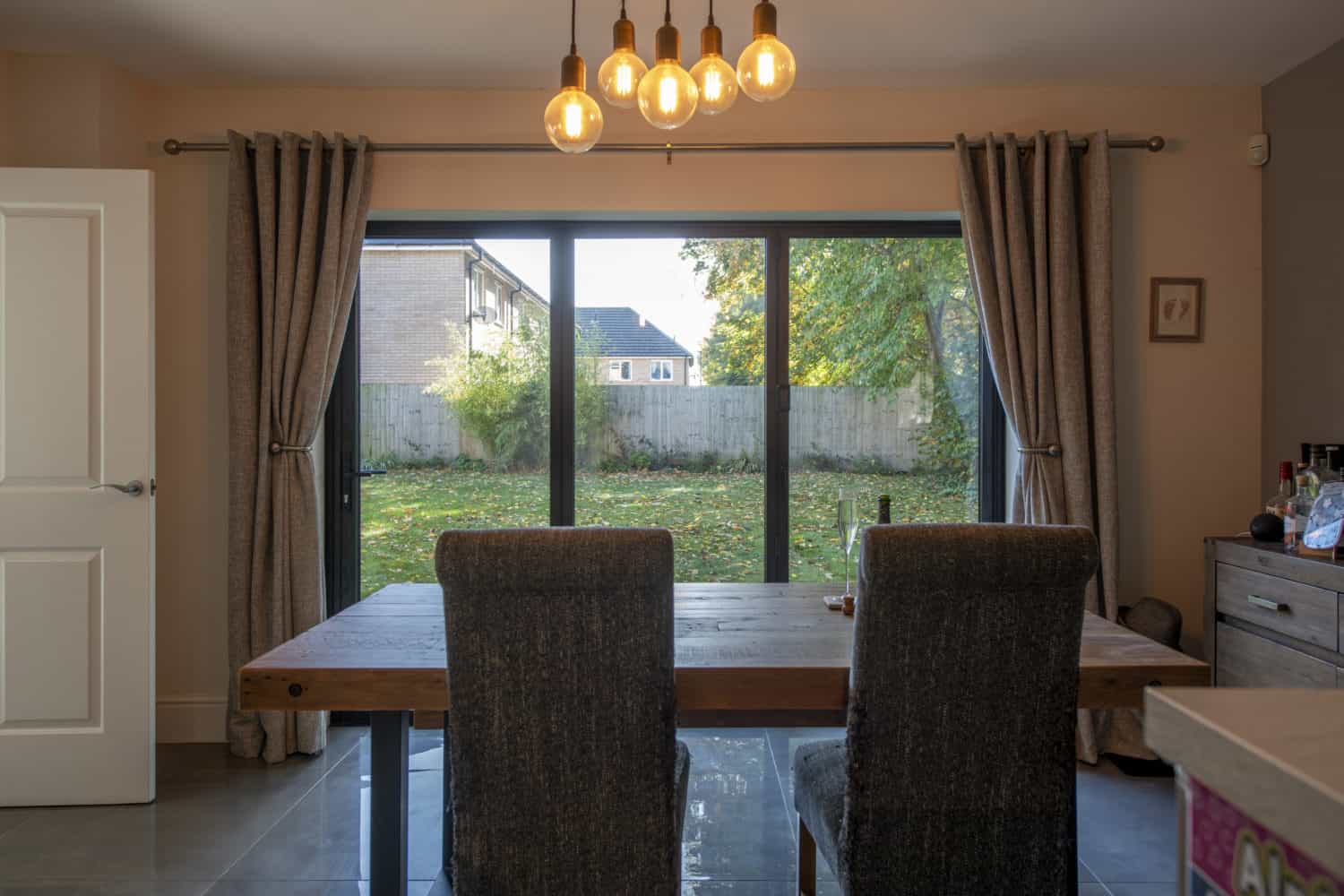 A dining room with a wooden table and two upholstered chairs faces large sliding doors that overlook a backyard with trees and a wooden fence. Warm pendant lights hang from the ceiling, creating a cozy atmosphere.