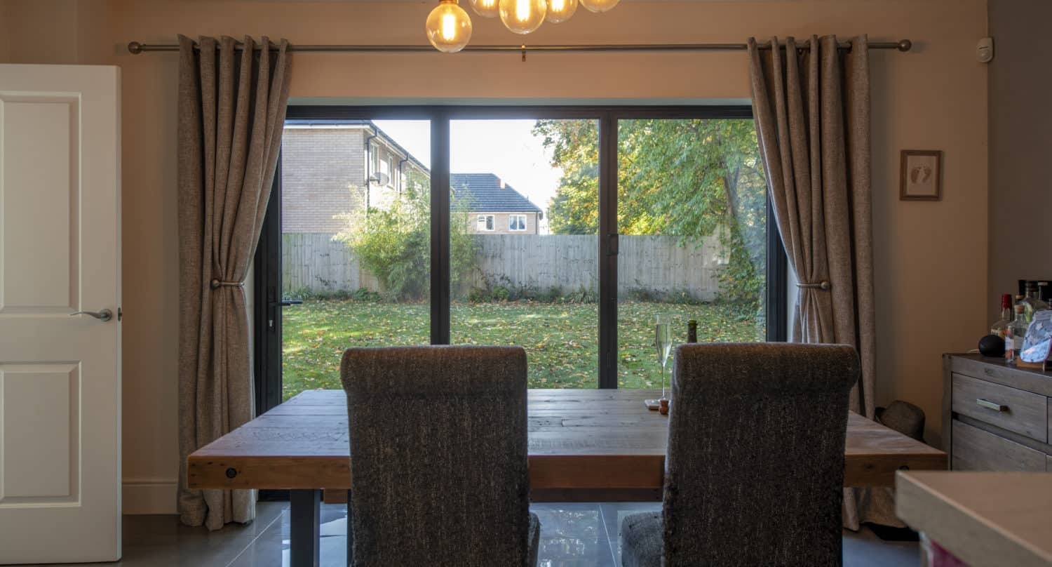A dining room with a wooden table and two upholstered chairs faces large sliding doors that overlook a backyard with trees and a wooden fence. Warm pendant lights hang from the ceiling, creating a cozy atmosphere.
