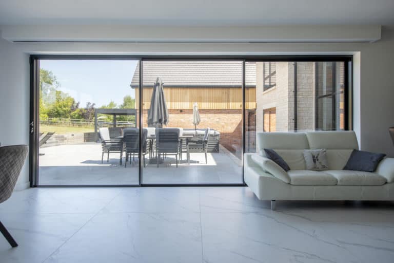 A modern living room with white marble flooring and a white sofa, featuring large bifold doors that open to a patio with a dining table, chairs, and umbrellas outside.