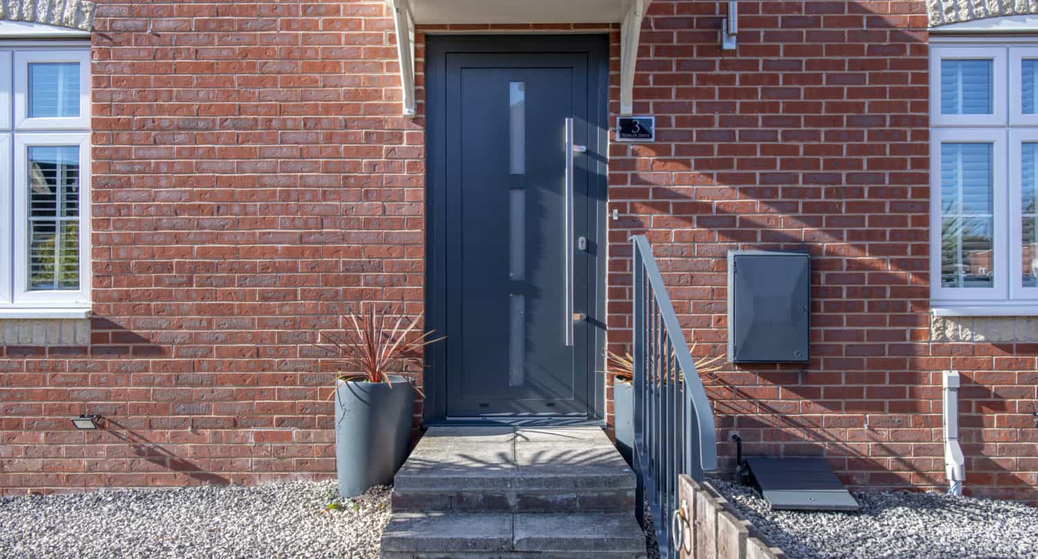 A modern dark gray front door with a metal handle, set in a red brick house featuring two windows, potted plants, and a mailbox. The entrance is enhanced by concrete steps and complemented by stylish bifold doors nearby.