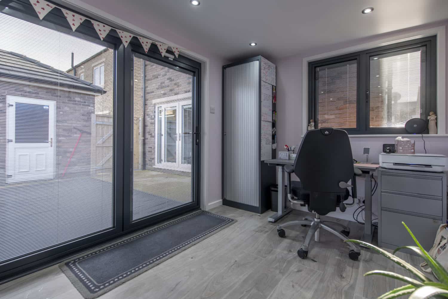 Modern home office with a black desk chair, grey desk, computer, and drawers by large bifold doors. Window with blinds, indoor plant, and outdoor area visible through the glass. Triangular bunting decorates the ceiling.