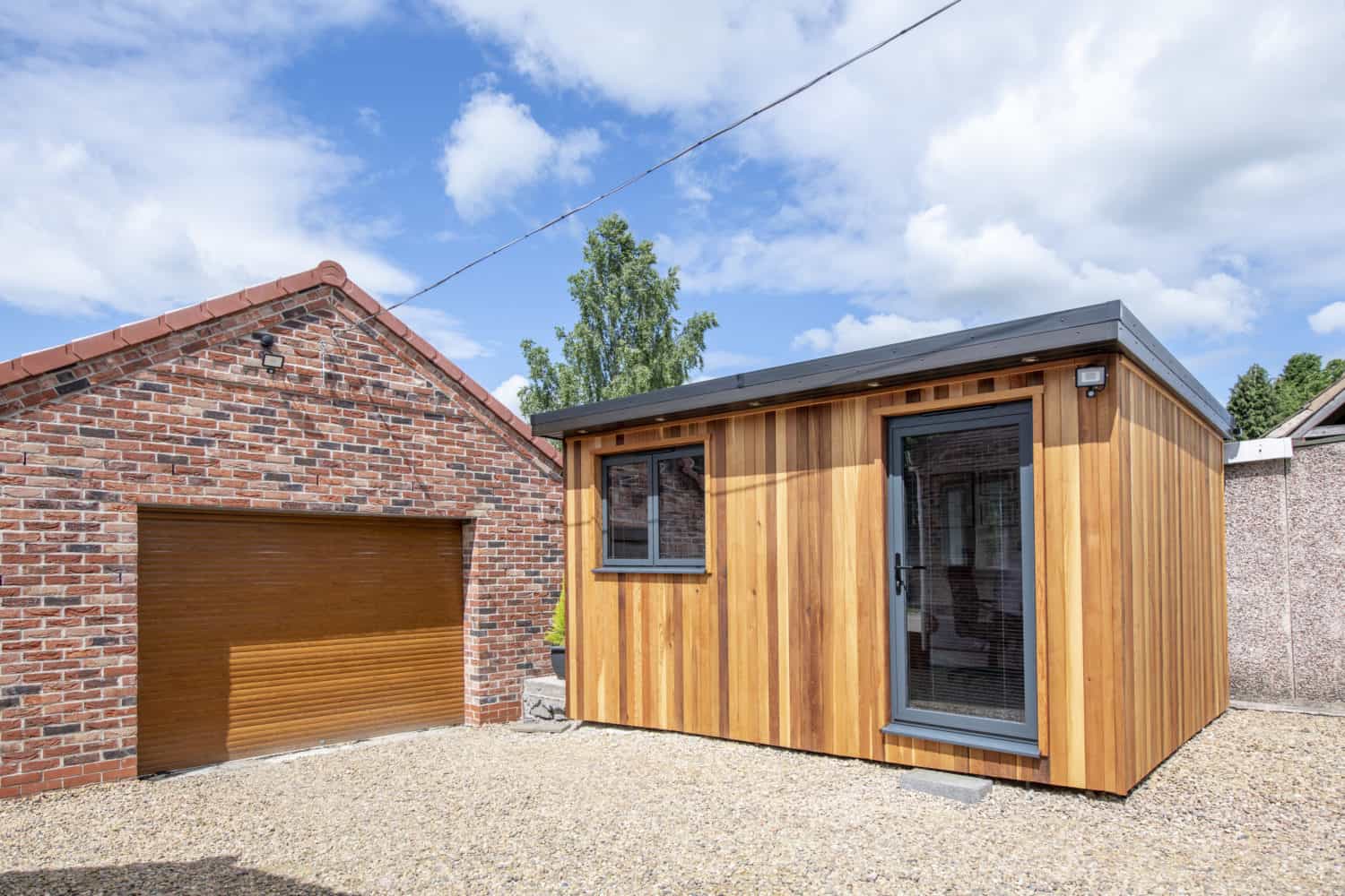 A small modern garden room with wooden siding and large windows stands next to a brick garage with a brown front door, set on a gravel driveway under a partly cloudy sky.