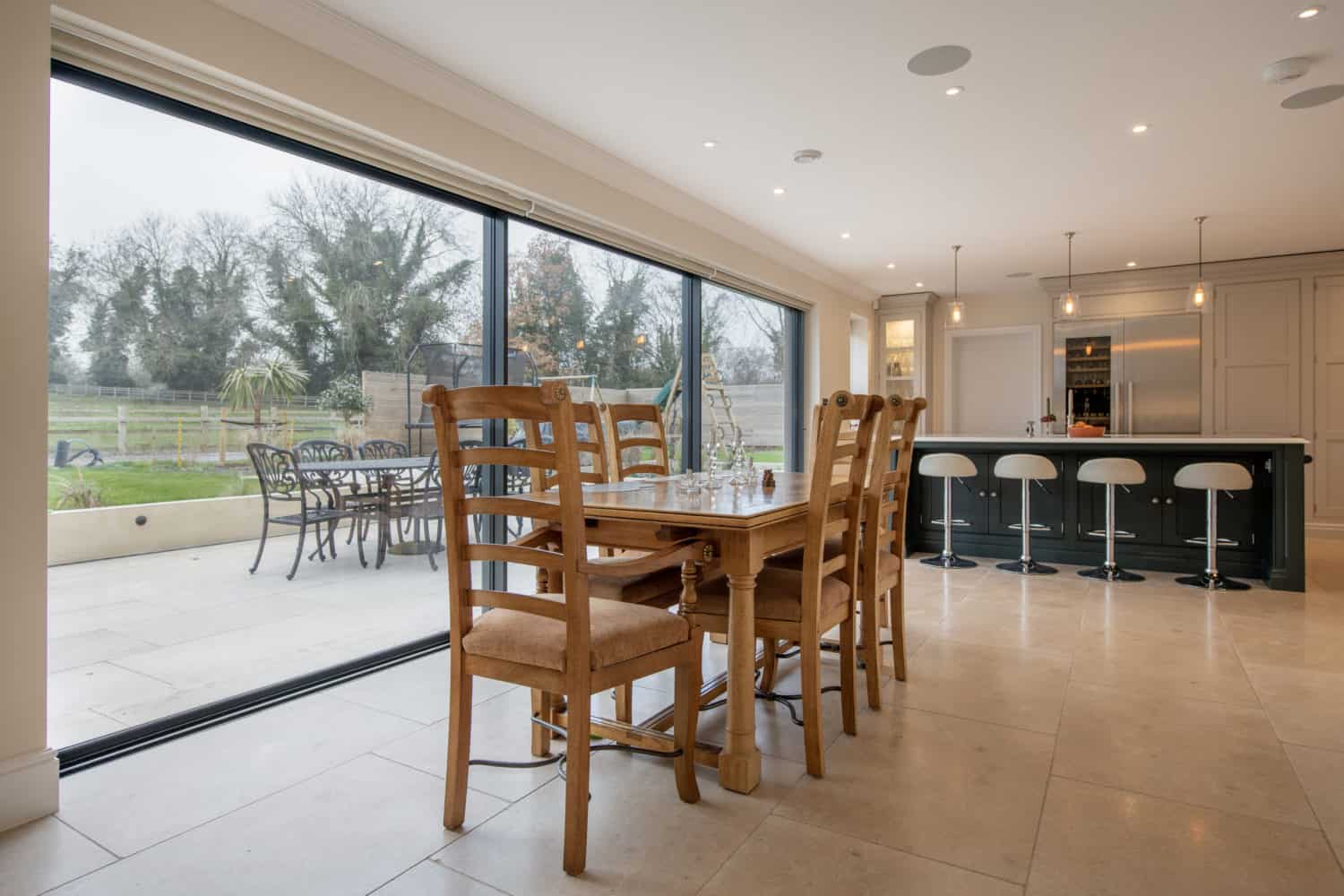 Modern kitchen and dining area with a wooden table and chairs near bespoke glass sliding doors, overlooking a patio with outdoor seating and a grassy yard with trees in the background.