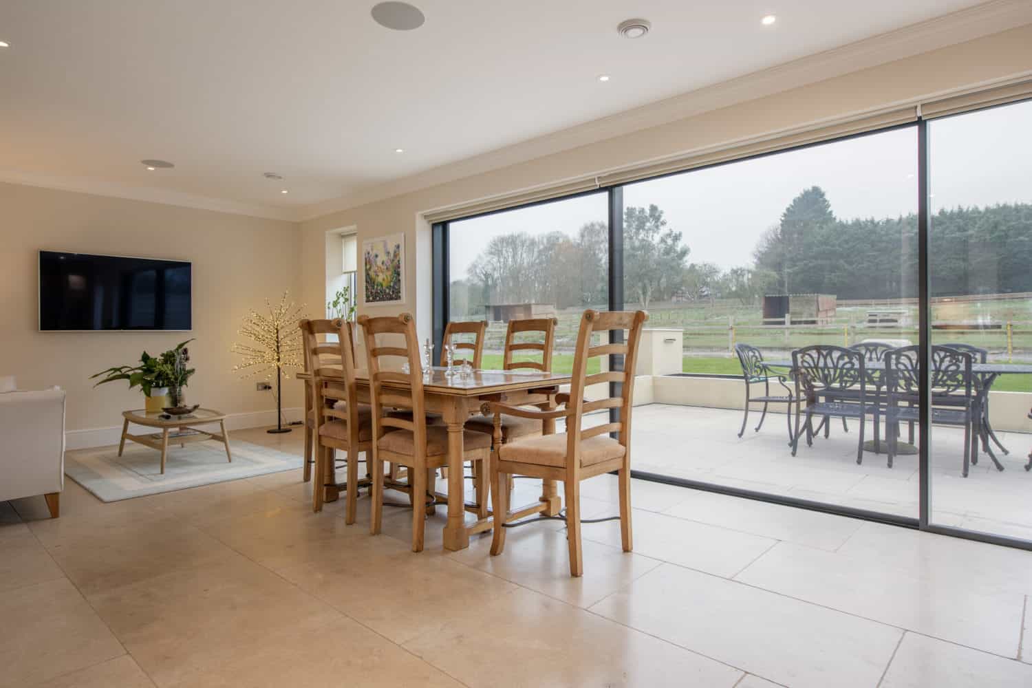 Modern dining area with a wooden table and chairs next to expansive bifold doors overlooking a patio with outdoor furniture and a scenic view of trees and fields.