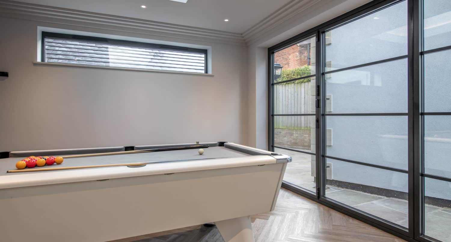 A modern room with a white pool table, neatly arranged balls, herringbone wood floor, large sliding doors, a skylight, and a horizontal window filling the space with natural light.