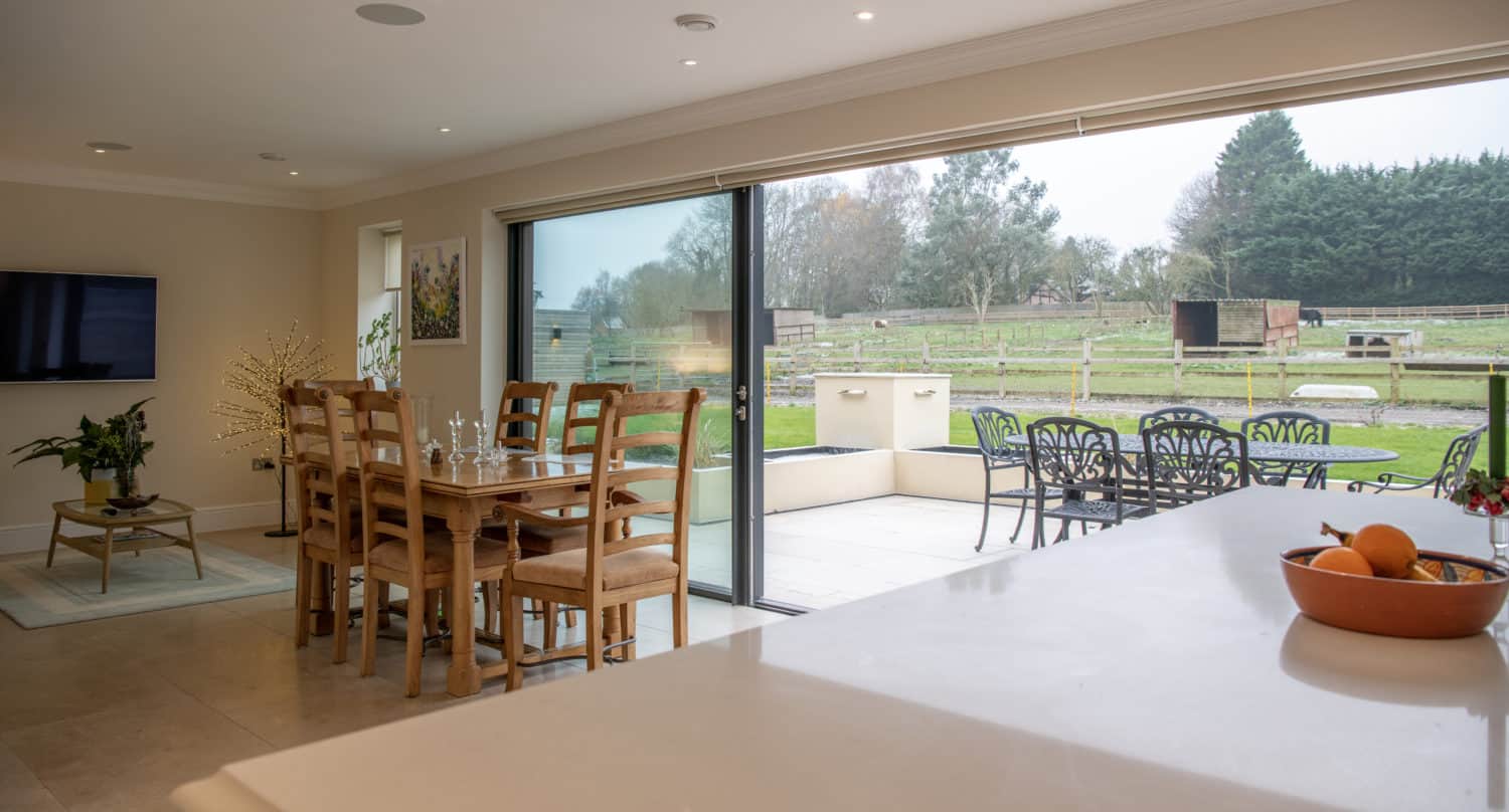 A modern open-plan dining and living area with wooden furniture, bespoke glass solutions in the sliding doors opening to a patio with outdoor seating, and a view of a green field and trees. A bowl of fruit sits on a white countertop.