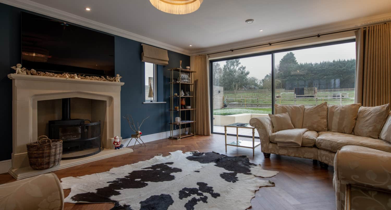 A cozy living room with a curved beige sofa, cowhide rug, fireplace with a wood-burning stove, large TV above the mantel, shelves, and bespoke sliding doors overlooking a garden and trees.