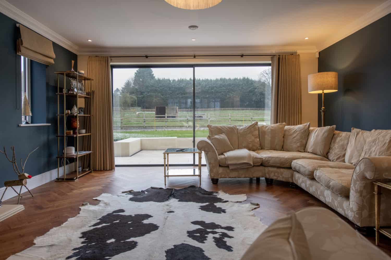 A modern living room with a beige sectional sofa, floor lamp, glass coffee table, and cowhide rug on wooden floors. Large sliding bifold doors offer a view of a green garden and trees outside.