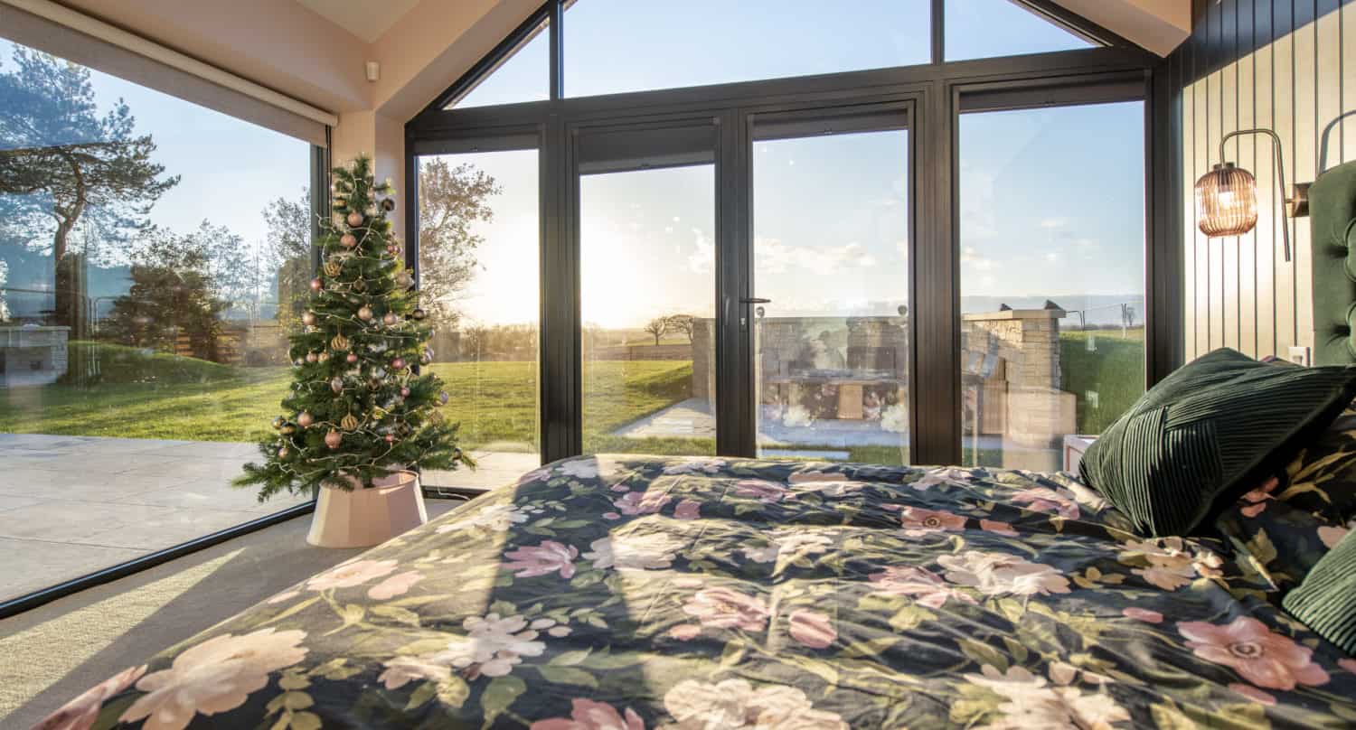A cozy bedroom with large floor-to-ceiling windows, a floral-patterned bedspread, and a decorated Christmas tree in the corner. Sunlight streams in through bifold doors, revealing a scenic outdoor view of trees and grass.
