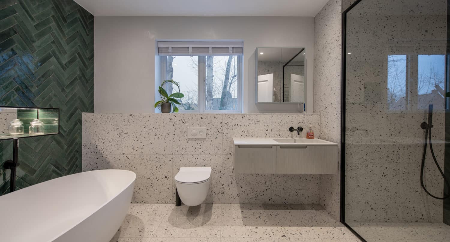 Modern bathroom with a freestanding tub, wall-mounted toilet, floating vanity, and a glass-enclosed shower featuring bespoke glass solutions. Green tile accent wall, terrazzo surfaces, and a plant by the window add style and freshness.