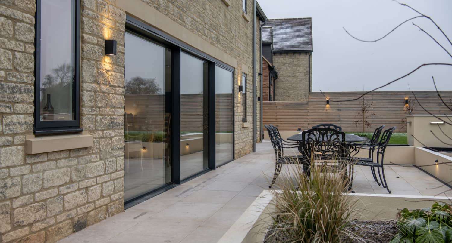 Modern stone house with large sliding doors leading to a patio. Outdoor dining set, decorative lighting, and landscaped plants are visible beneath the elegant glass roofing in the patio area.